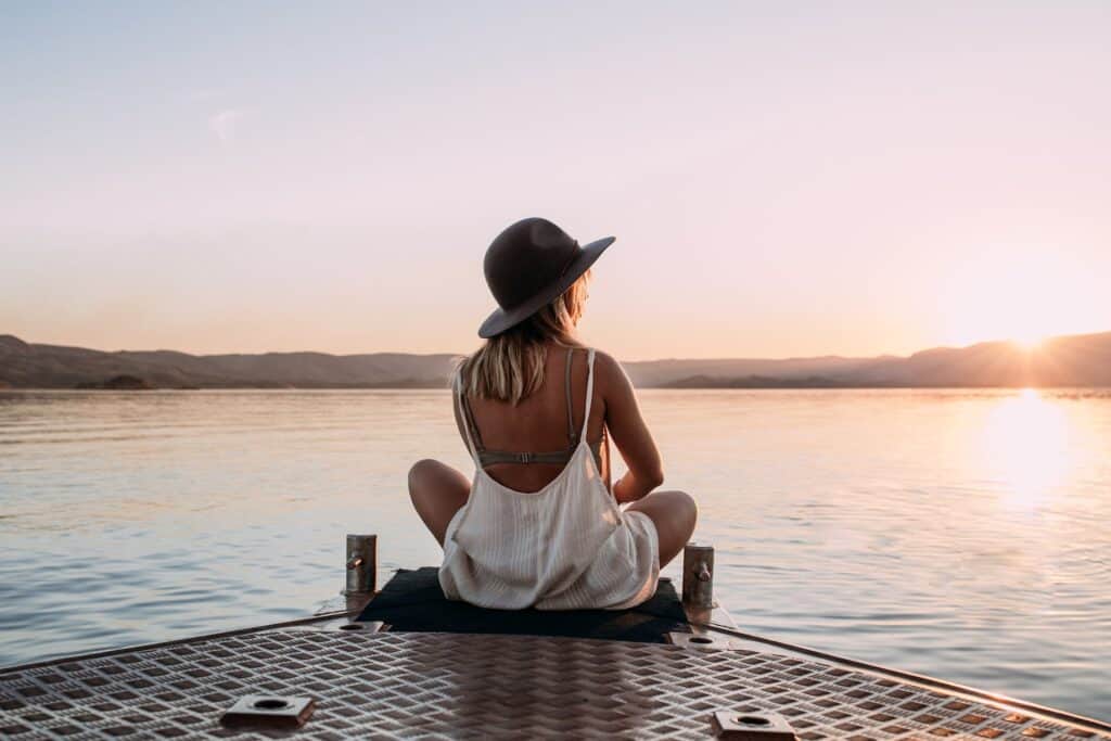 Woman in loose white clothing and a hat sitting on the end of a dock looking at a lake.